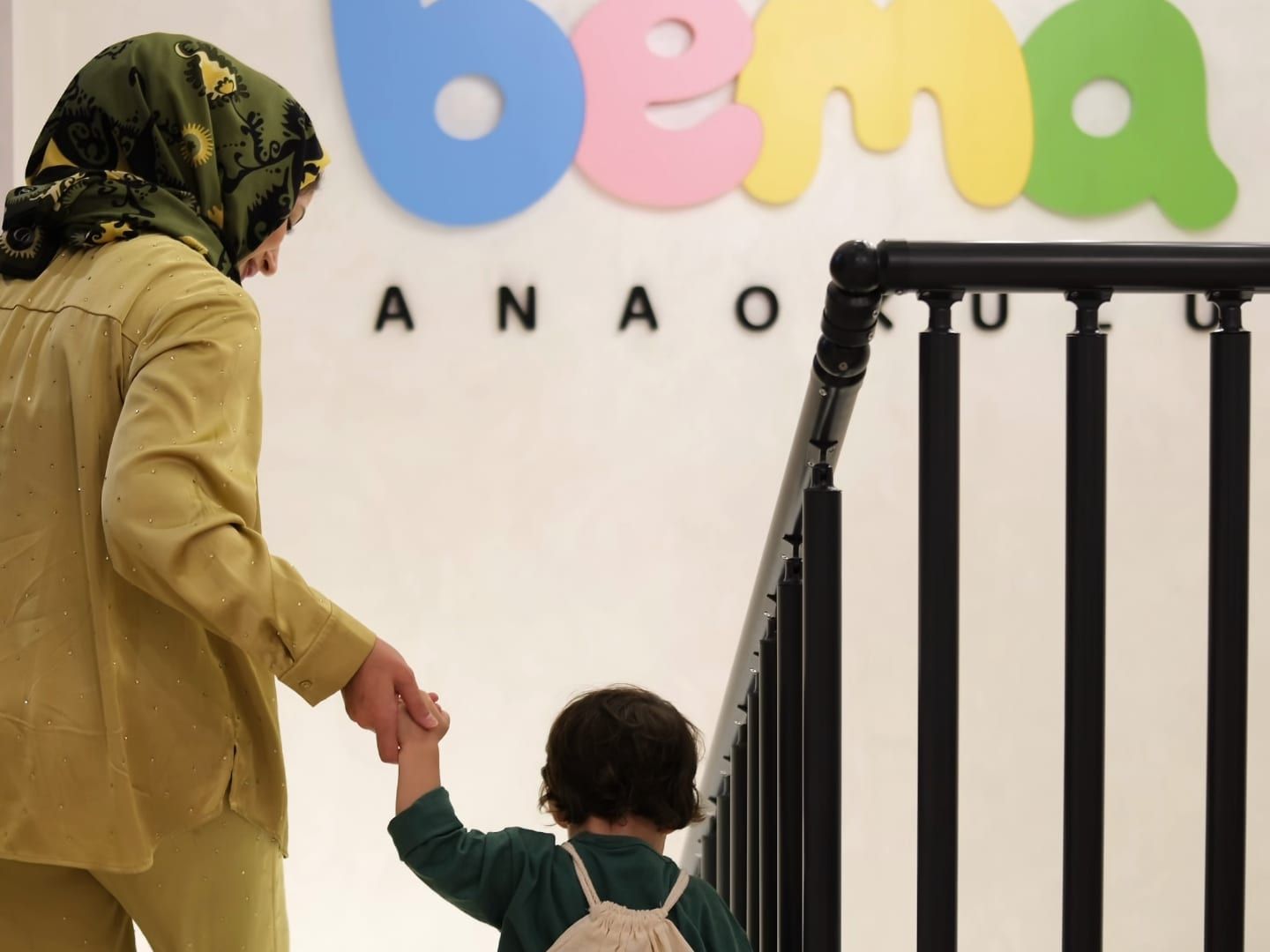 BEMA logolu renkli duvarın önünde el ele merdivene yönelen veli ve çocuk, arkadan